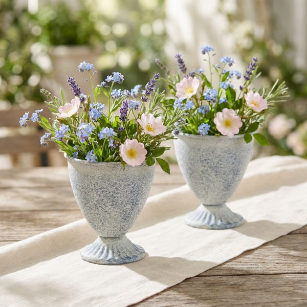 Zwei dekorative Pflanzgefäße in Kelchform auf einem Tisch mit sommerlichen Blumenarrangements.