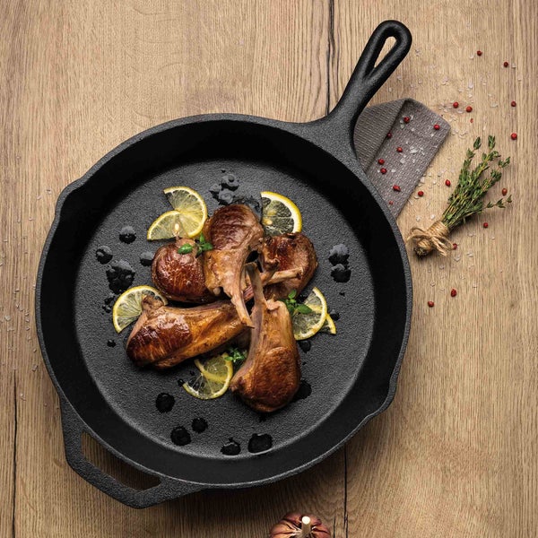 Schwarze gusseiserne Pfanne mit Lammkoteletts und Zitronenscheiben auf einem Holztisch.
