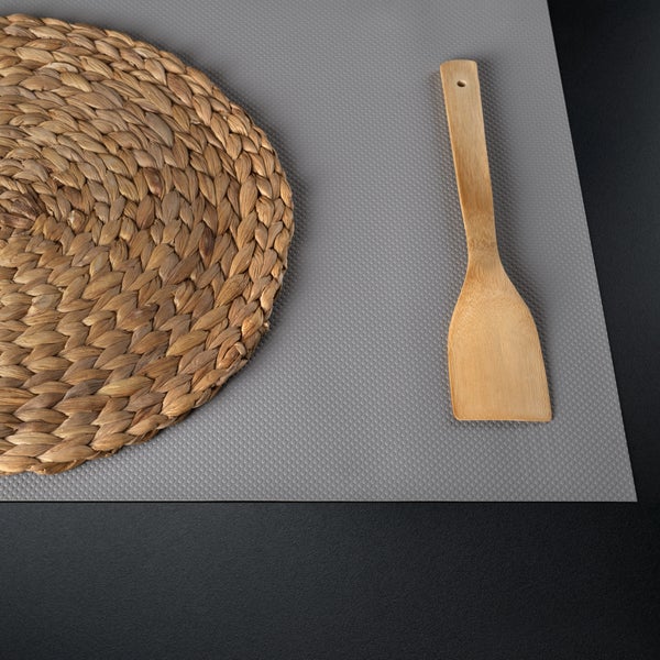 Strukturierte graue Antirutschmatte für Schubladen mit einem runden geflochtenen Platzset und einem Pfannenwender aus Holz.