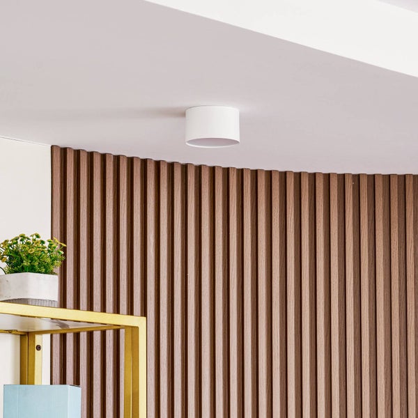 Moderner weißer zylindrischer Deckenstrahler an einer weißen Decke neben einer Wand mit braunen vertikalen Holzlamellen.