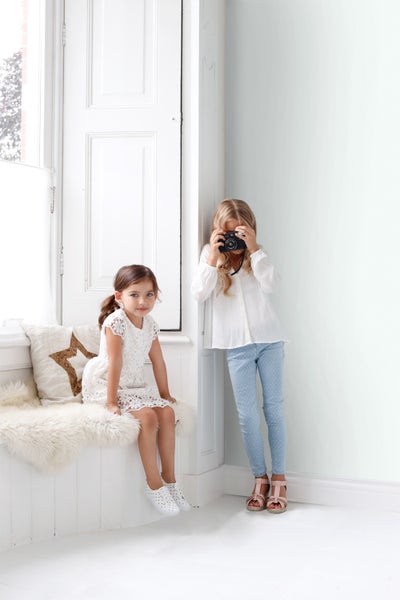 Zwei Mädchen in einem hellen Raum, eines mit einer Kamera, das andere sitzt auf einer Fensterbank