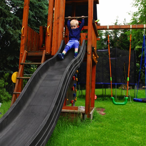 Spielturm aus Holz im Garten mit Rutsche, Kletterwand und Schaukel, ein Kind rutscht auf der dunklen Kunststoffrutsche.