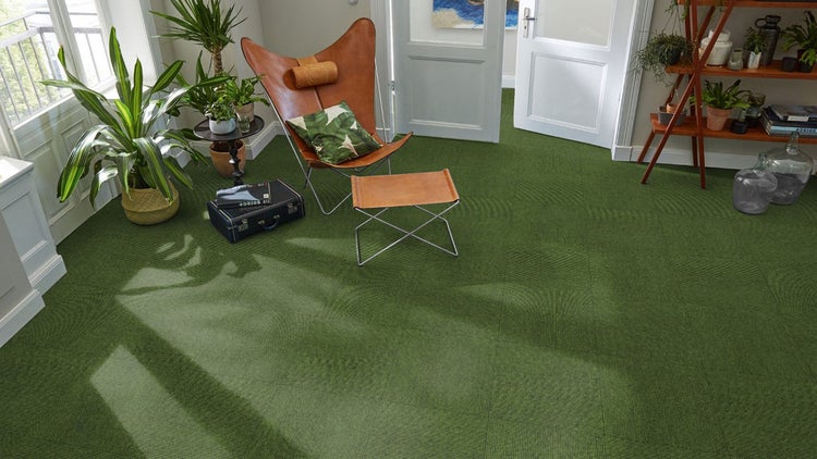 Grüne Teppichfliesen in einem hellen Wohnraum mit Butterfly-Sessel, Zimmerpflanzen und einem Holzregal.