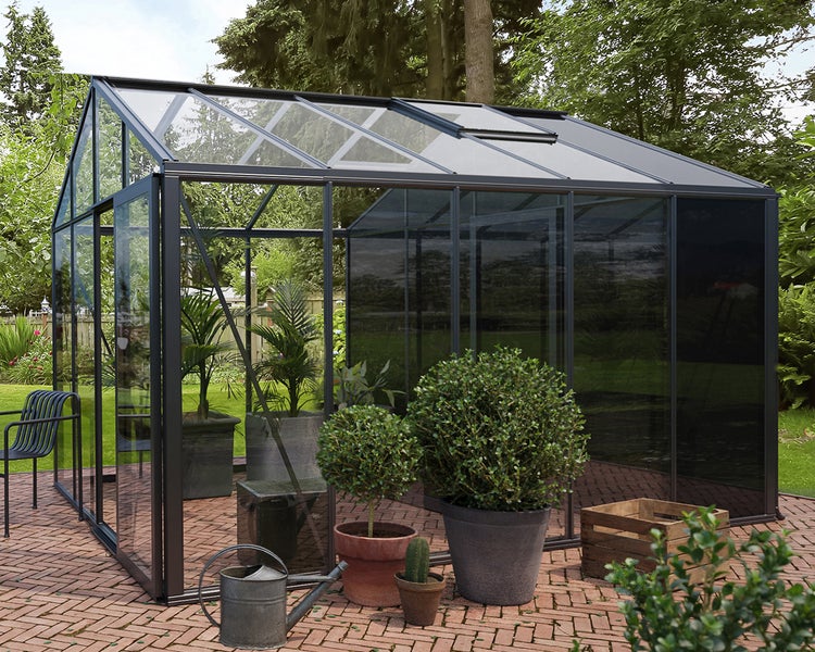 Modernes Gewächshaus aus Glas mit dunklem Metallrahmen auf einer gepflasterten Terrasse im Garten, hell und organisiert mit verschiedenen Topfpflanzen.