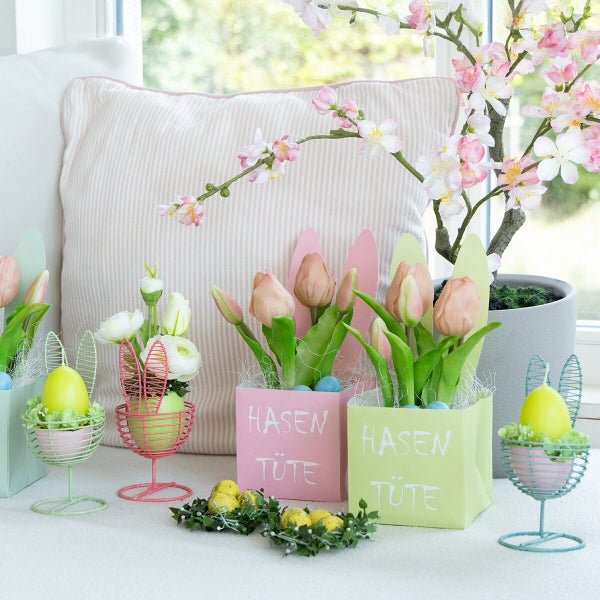 Osterdekoration mit Hasentüten aus Papier, Drahteierbechern und künstlichen Blumenarrangements vor einem Fenster.