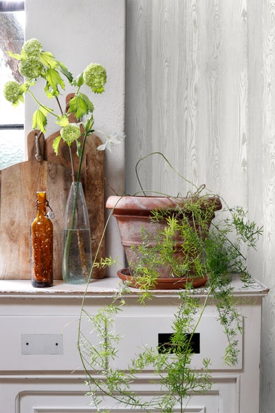 Dekoratives Arrangement mit Blumen, Flaschen und Topfpflanze auf einem Schrank vor einer Wandtapete in Holzoptik.