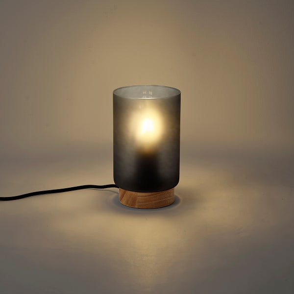 Moderne Tischleuchte mit zylindrischem Glasschirm und Holzsockel leuchtet warm.