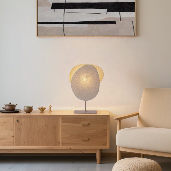 Moderne Tischleuchte mit organisch geformtem Textilschirm auf einem Sideboard aus Holz in einem hellen Wohnzimmer.