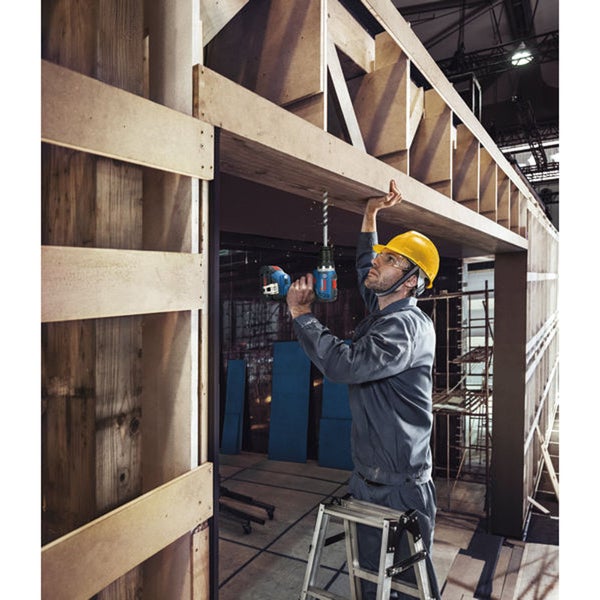 Ein Mann bohrt mit einer Bohrmaschine in ein Holzgerüst, während er auf einer Leiter steht und einen Schutzhelm trägt.