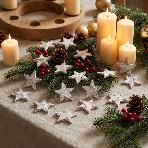 Weihnachtliche Tischdekoration mit ausgestreuten Holzsternen, Tannenzweigen, Zapfen, Beeren und brennenden Stumpenkerzen auf einer Leinentischdecke.