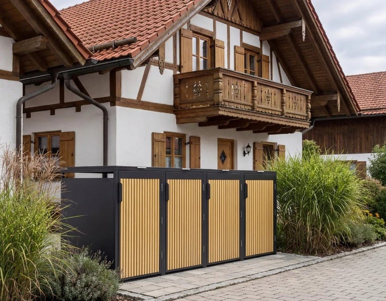 Moderne Mülltonnenbox für vier Tonnen aus Metall mit Holzlatten vor einem Wohnhaus in einem gepflegten Außenbereich.