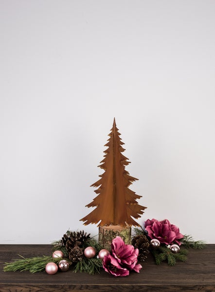 Dekorativer Weihnachtsbaum aus Metall mit Rost-Patina auf einem Holzsockel, umgeben von Tannenzweigen, Zapfen und rosa Weihnachtsdekorationen.