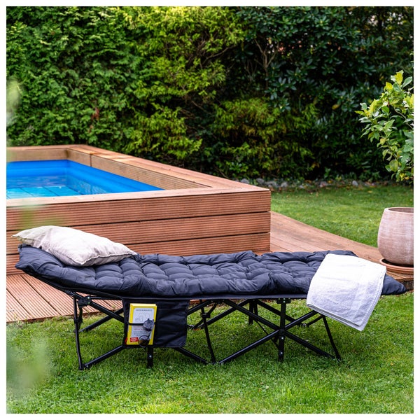 Schwarze klappbare Gartenliege mit gepolsterter Auflage, Kissen und Handtuch auf einer Rasenfläche neben einer hölzernen Pooleinfassung.
