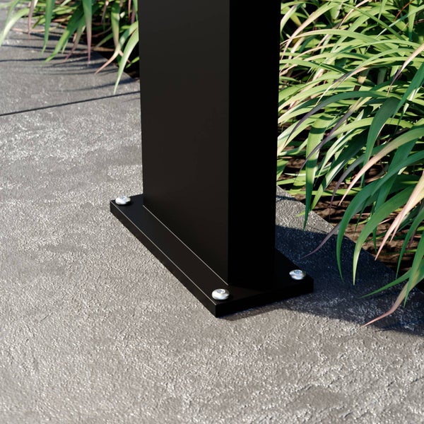 Basis eines schwarzen Metallpfostens mit Bodenplatte, befestigt mit silbernen Schrauben auf grauem Untergrund neben grünen Pflanzen.