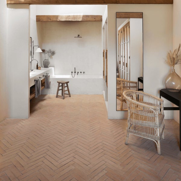 Badezimmer mit hellen Wänden, Holzbalken an der Decke und Bodenfliesen in Fischgrätoptik, ergänzt durch eine Badewanne und einen Rattanstuhl.