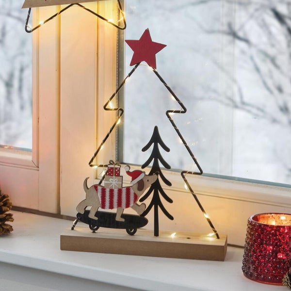 Weihnachtliche Holzdekoration mit LED Beleuchtung in Form eines Tannenbaums mit Dackel auf einem Skateboard auf einer Fensterbank.