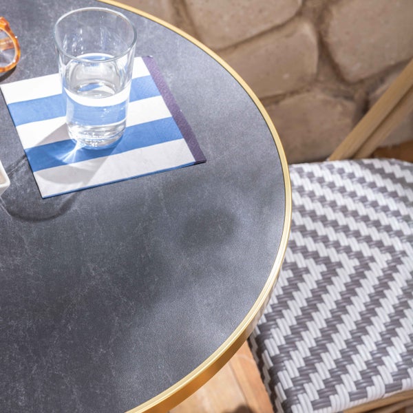 Runder Bistrotisch mit dunkler Tischplatte in Steinoptik und goldfarbenem Metallrand, kombiniert mit einem Flechtstuhl.