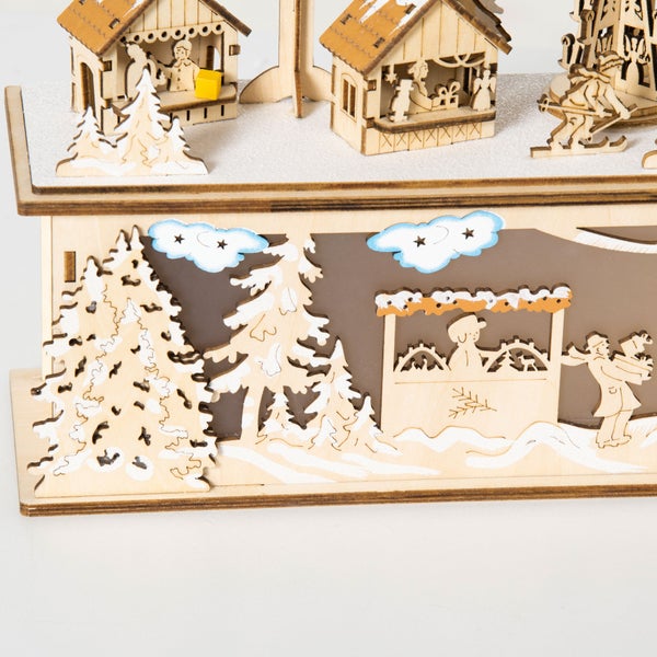 Detailansicht einer hölzernen Weihnachtsdekoration mit verschneiter Dorfszene, kleinen Häusern und Winterbäumen.