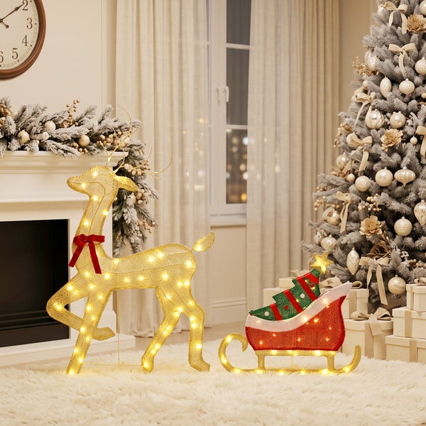 Beleuchtete Weihnachtsdekoration Rentier mit Schlitten vor einem geschmückten Weihnachtsbaum und Kamin in einem weihnachtlich dekorierten Wohnzimmer.