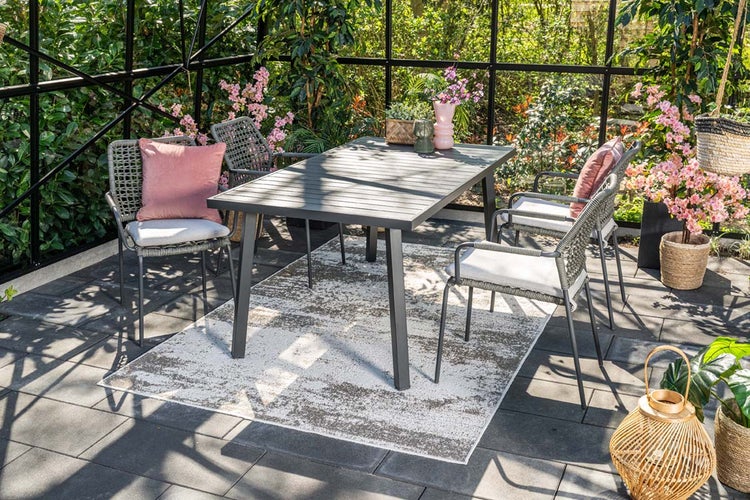 Moderne Garten-Essgruppe mit grauem Lattentisch und vier Seilgeflecht-Stühlen mit Auflagen auf einem Outdoorteppich in einem hellen Wintergarten.