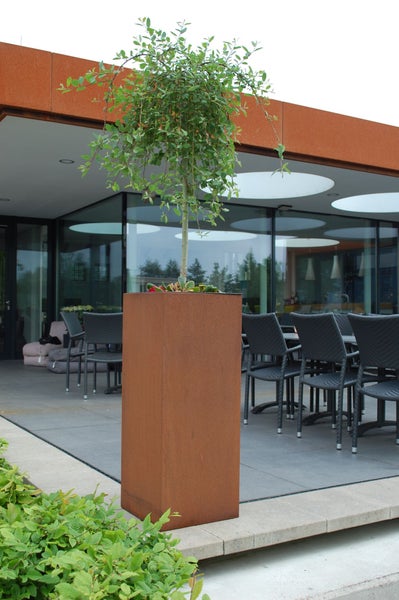 Hoher, quadratischer Pflanzkübel in Edelrost-Optik mit einem kleinen Baum auf einer modernen Terrasse mit Gartenstühlen.