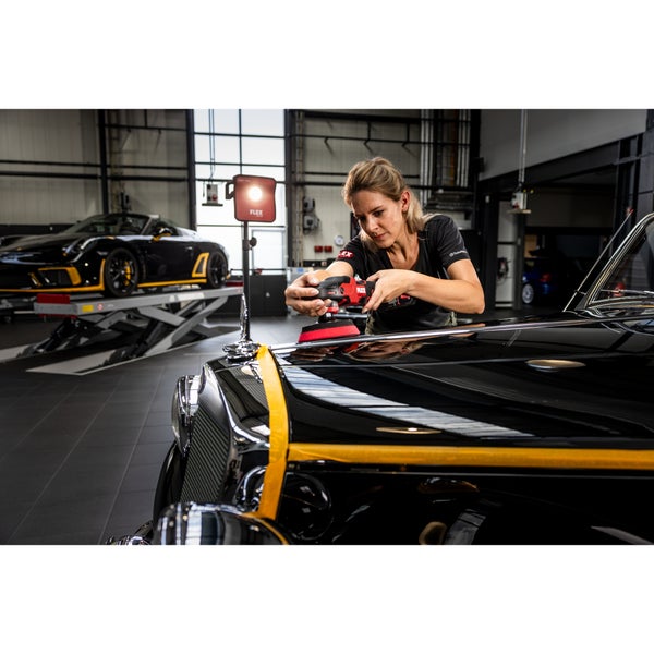 Eine Frau poliert die Motorhaube eines Oldtimers mit einer Poliermaschine in einer professionellen Werkstatt mit einem Stativstrahler.