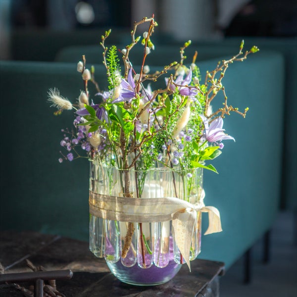 Frühlingsgesteck mit lila Blüten und Weidenkätzchen in einer kreisförmigen Vase aus Glasröhrchen, die mit einer hellen Schleife gebunden ist.
