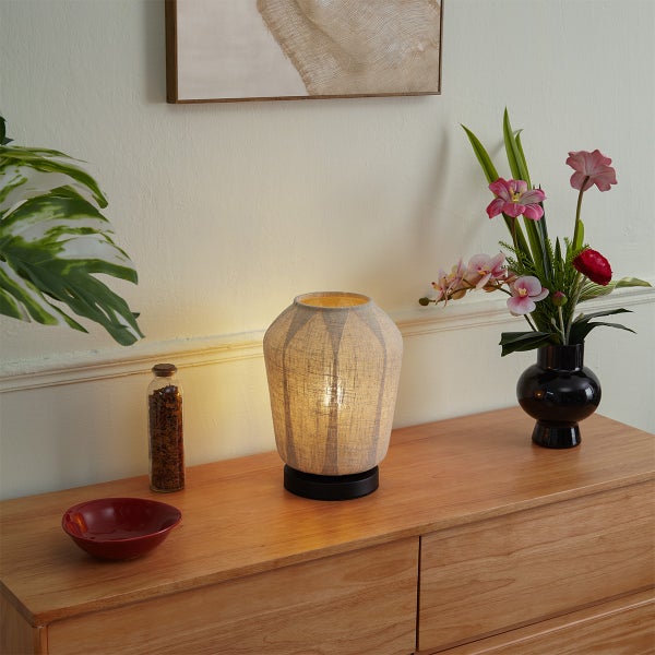 Moderne Tischleuchte mit beigem Textilschirm und schwarzem Fuß auf einem Holzsideboard, neben Blumenvase, roter Schale und dekorativer Flasche.