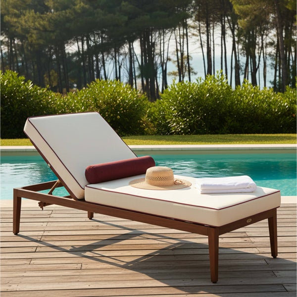 Sonnenliege aus Holz mit weißer Auflage und rotem Kissen auf einer Holzterrasse neben einem Swimmingpool.