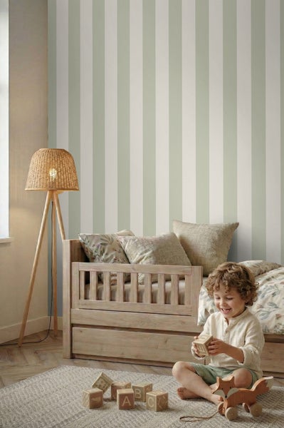 Helles Kinderzimmer mit gestreifter Tapete, einem Holzbett, einer Rattan-Stehlampe und einem Kind, das mit Holzspielzeug auf einem Teppich spielt.