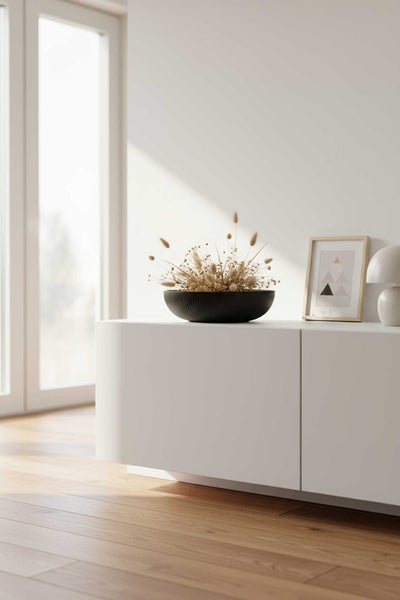 Minimalistisches weißes Sideboard mit einer schwarzen Dekoschale mit Trockenblumen, einem Bilderrahmen und einer Lampe in einem hellen Raum.