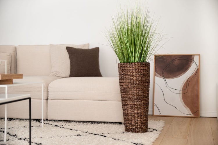 Künstliches Ziergras in einem hohen, geflochtenen Übertopf in einem hellen, modern eingerichteten Wohnzimmer neben einem Sofa.