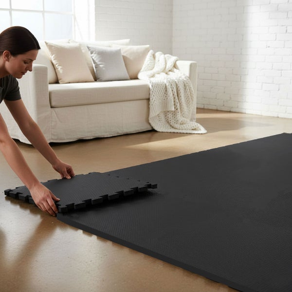 Eine Person setzt schwarze Puzzlematten aus Schaumstoff in einem hellen Wohnzimmer mit weißem Sofa und Ziegelwand zusammen.