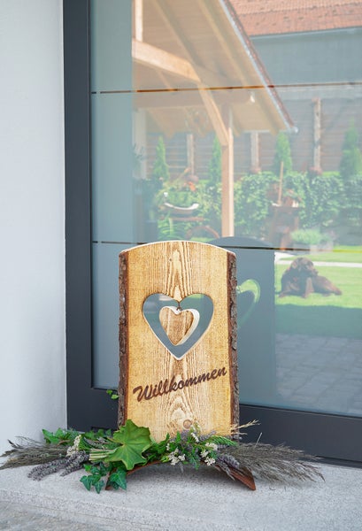 Dekoratives Holzschild mit Willkommensschriftzug und Herzausschnitt vor einer Glasfront mit Gartenblick
