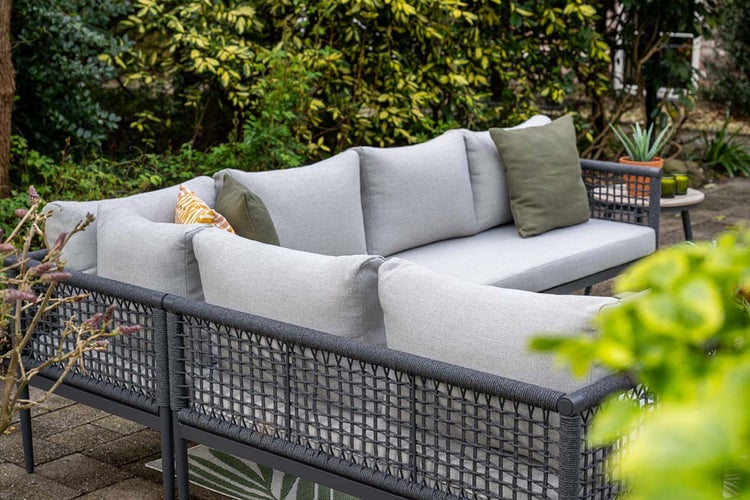 Moderne Garten-Lounge-Ecksofa mit dunklem Geflechtrahmen, hellgrauen Sitzkissen und Zierkissen in einem grünen Garten.