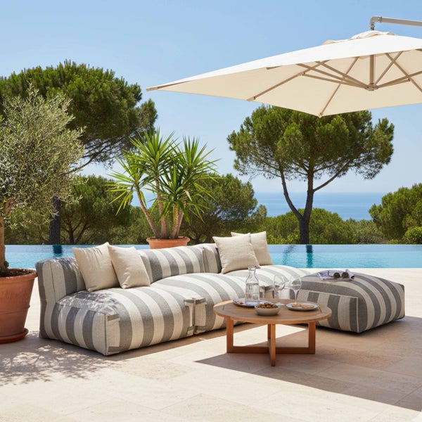 Gestreiftes Outdoor-Lounge-Set mit Sofa und Hocker auf einer Terrasse am Pool, runder Holztisch, Sonnenschirm und Blick auf das Meer.