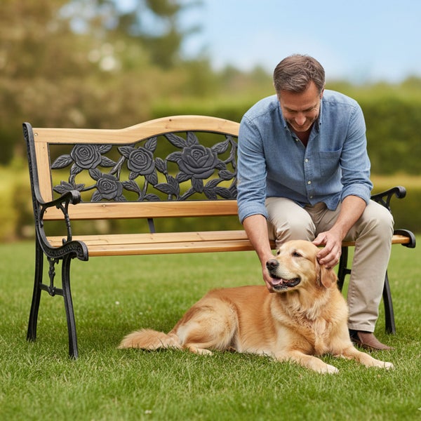Gartenbank aus Holz und Gusseisen mit dekorativem Rosenmuster auf einer Rasenfläche mit einem Mann und einem Hund.