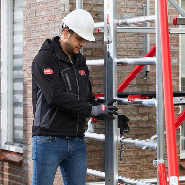 Mann mit Schutzhelm und Altrex Jacke montiert ein Altrex Gerüst vor einer Backsteinwand