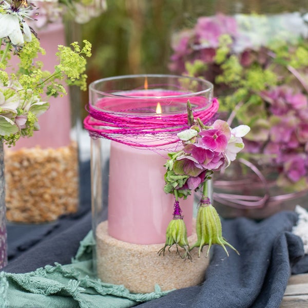 Dekorative Glasvase mit brennender Stumpenkerze auf Sand, verziert mit Kordel und Trockenblumen in einer hellen Gartenumgebung.