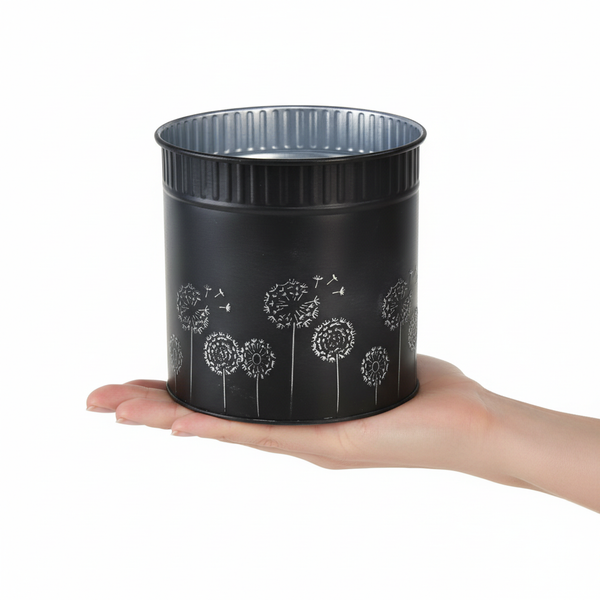 Schwarzer Übertopf aus Metall mit Pusteblumen-Muster, zur Größenveranschaulichung in einer Hand gehalten.
