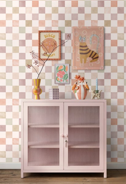 Eklektischer Wohnbereich mit rosa Metallschrank mit Riffelglastüren vor pastellfarben karierter Tapete, dekoriert mit bunten Vasen und Wandbildern.
