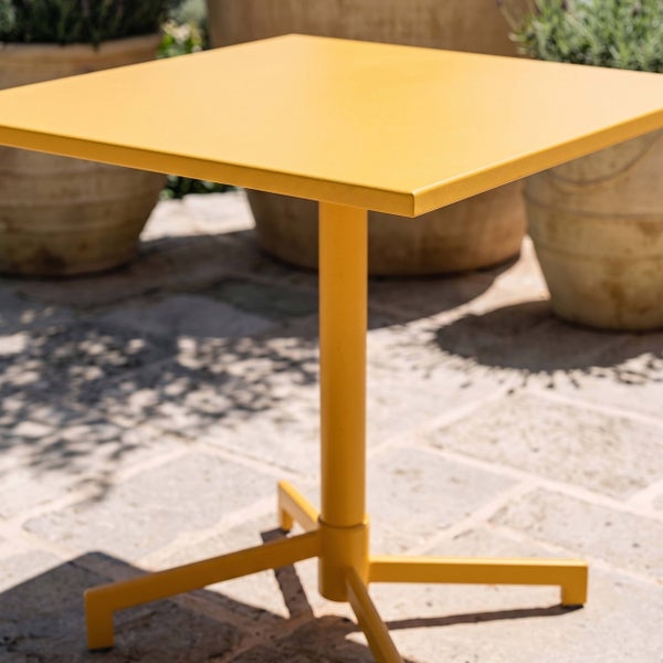 Gelber, quadratischer Gartentisch aus Metall mit einem Säulenfuß auf einer hellen, organisierten Steinterrasse.