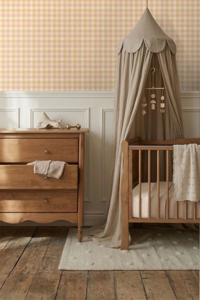Kinderzimmer mit Holzkommode und Babybett unter einem Stoffbaldachin. Im Hintergrund karierte Tapete und weiße Wandpaneele.
