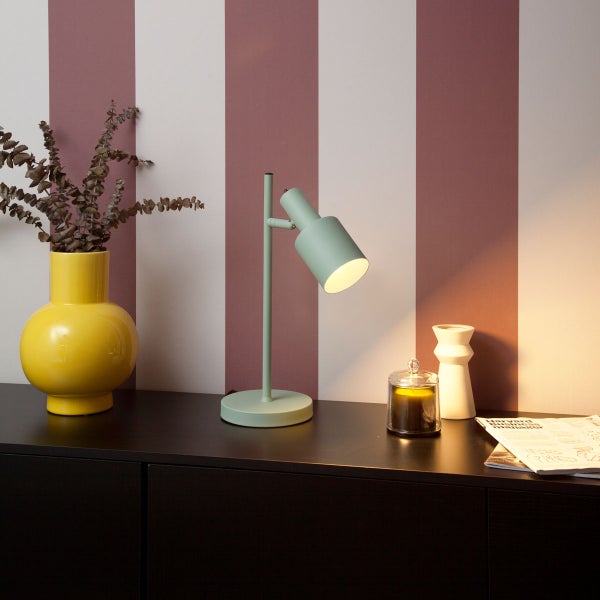 Mintgrüne Tischleuchte auf einem dunklen Sideboard vor einer gestreiften Wand mit einer gelben Vase, einer Kerze und einer Zeitschrift.