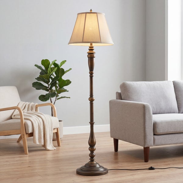 Klassische Stehleuchte in Bronzeoptik mit Stoffschirm in einem hellen Wohnzimmer neben Sofa und Sessel.