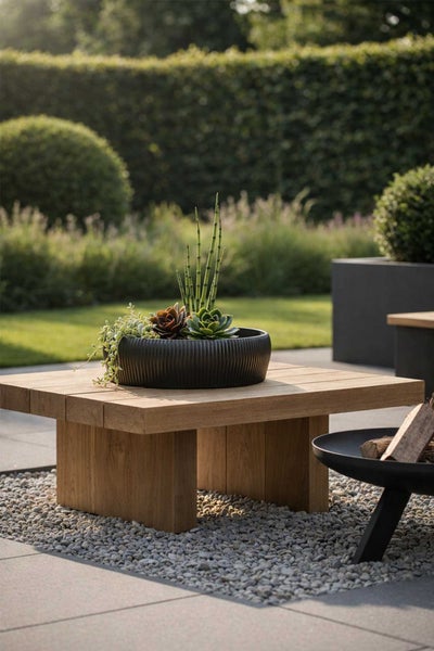 Quadratischer Gartentisch aus Holz mit schwarzer Pflanzschale und Sukkulenten in einem hellen, organisierten Garten auf Kies.