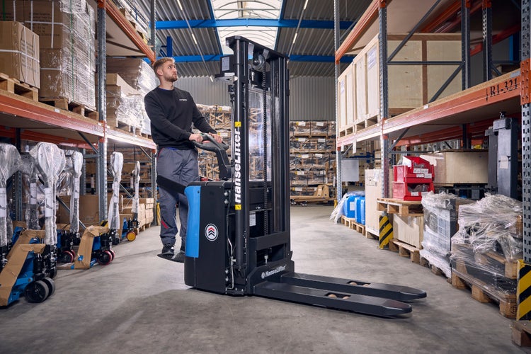 Ein Mitarbeiter bedient einen elektrischen Hochhubwagen von HanseLifter in einem Lagerhaus zwischen Regalen mit Paletten.