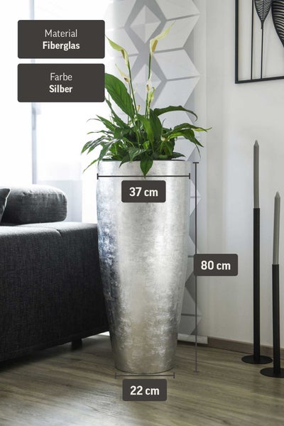 Hell eingerichtetes, modernes Wohnzimmer mit silberfarbener Bodenvase mit Kunstgras, dunklem Sofa und schlanken Kerzenständern.
