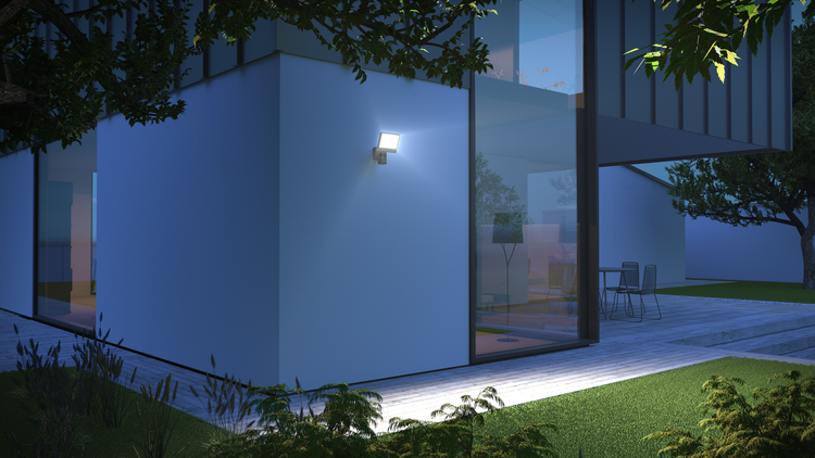 Moderner LED-Außenstrahler mit Bewegungsmelder an einer Hauswand montiert, beleuchtet nachts eine Terrasse aus Holz.