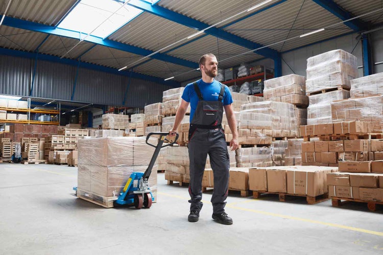 Ein Mann in Arbeitskleidung zieht einen beladenen Hubwagen durch ein großes Lager mit gestapelten Kartons auf Paletten.